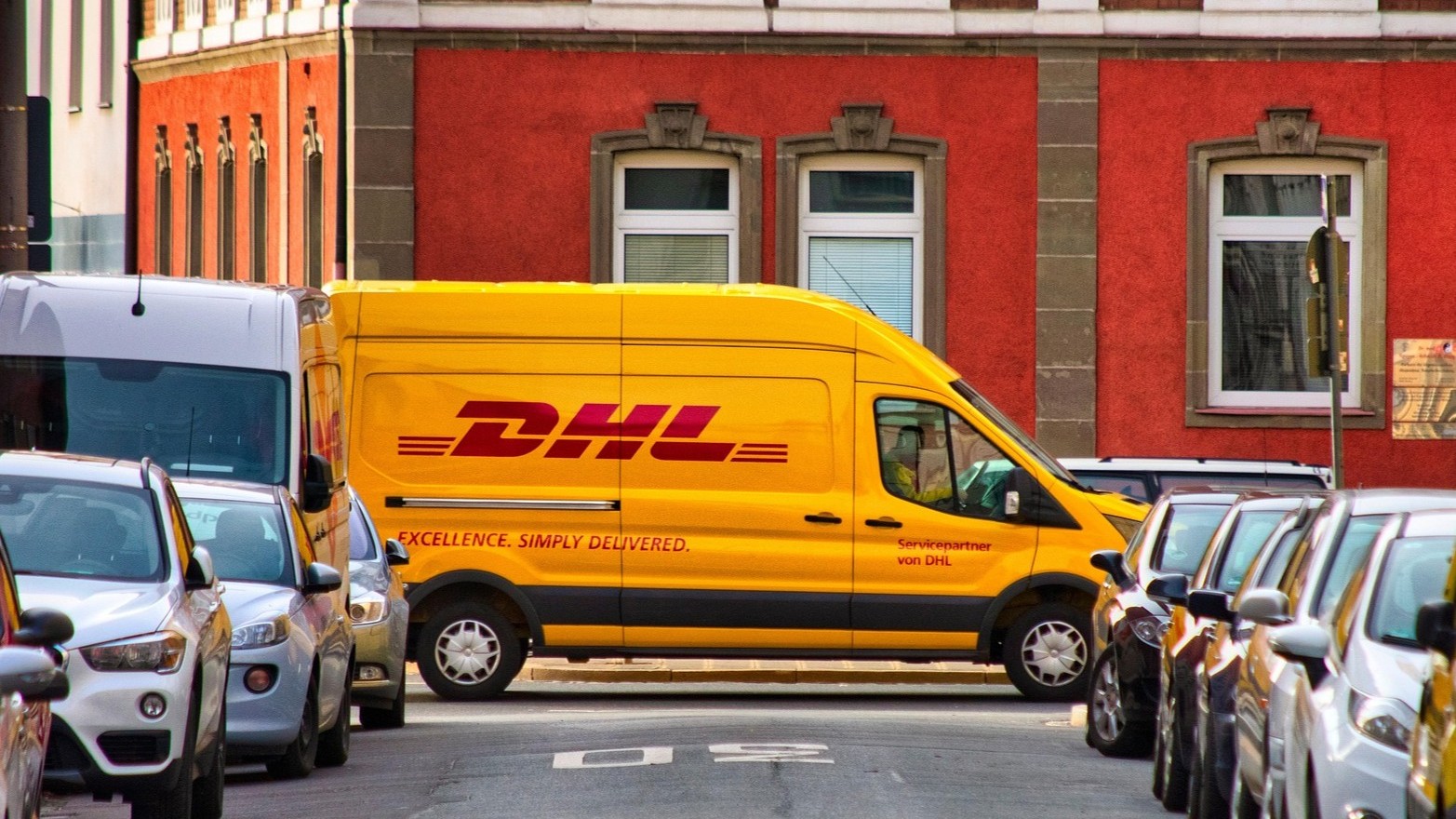 Foto eines gelben DHL-Lieferwagens, der auf einer städtischen Straße an parkenden Autos und einem roten Gebäude vorbeifährt.