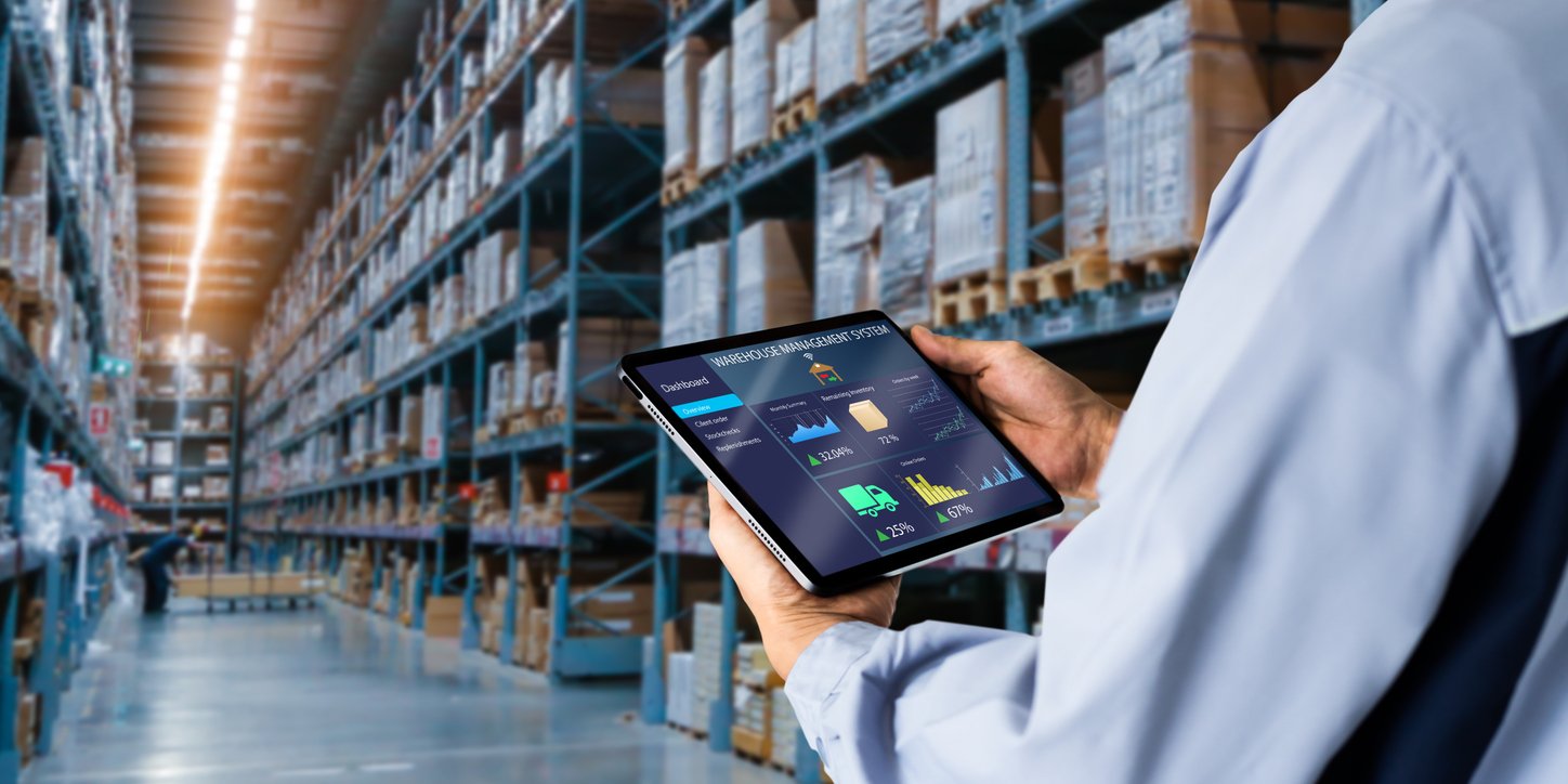 Bild einer Lagerhalle. Im Vordergrund ist ein Mann mit Tablet, auf dem eine Logistiksoftware geöffnet ist.