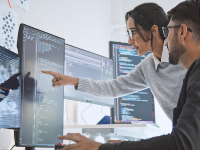 Eine Frau und ein Mann sitzen vor Computer-Bildschirmen und besprechen etwas.